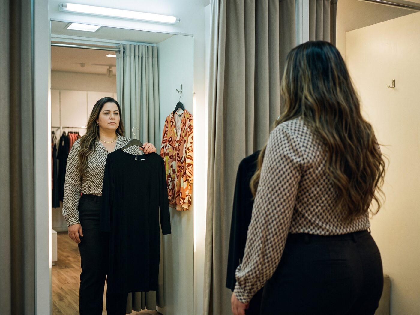 Mulher num provador segurando um vestido preto, com a peça estampada que ela tinha escolhido pendurada atrás